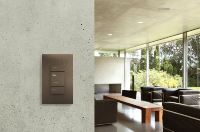 A wall-mounted keypad with labeled buttons for controlling various lighting and shading scenes, set against a concrete wall with a modern living room in the background.