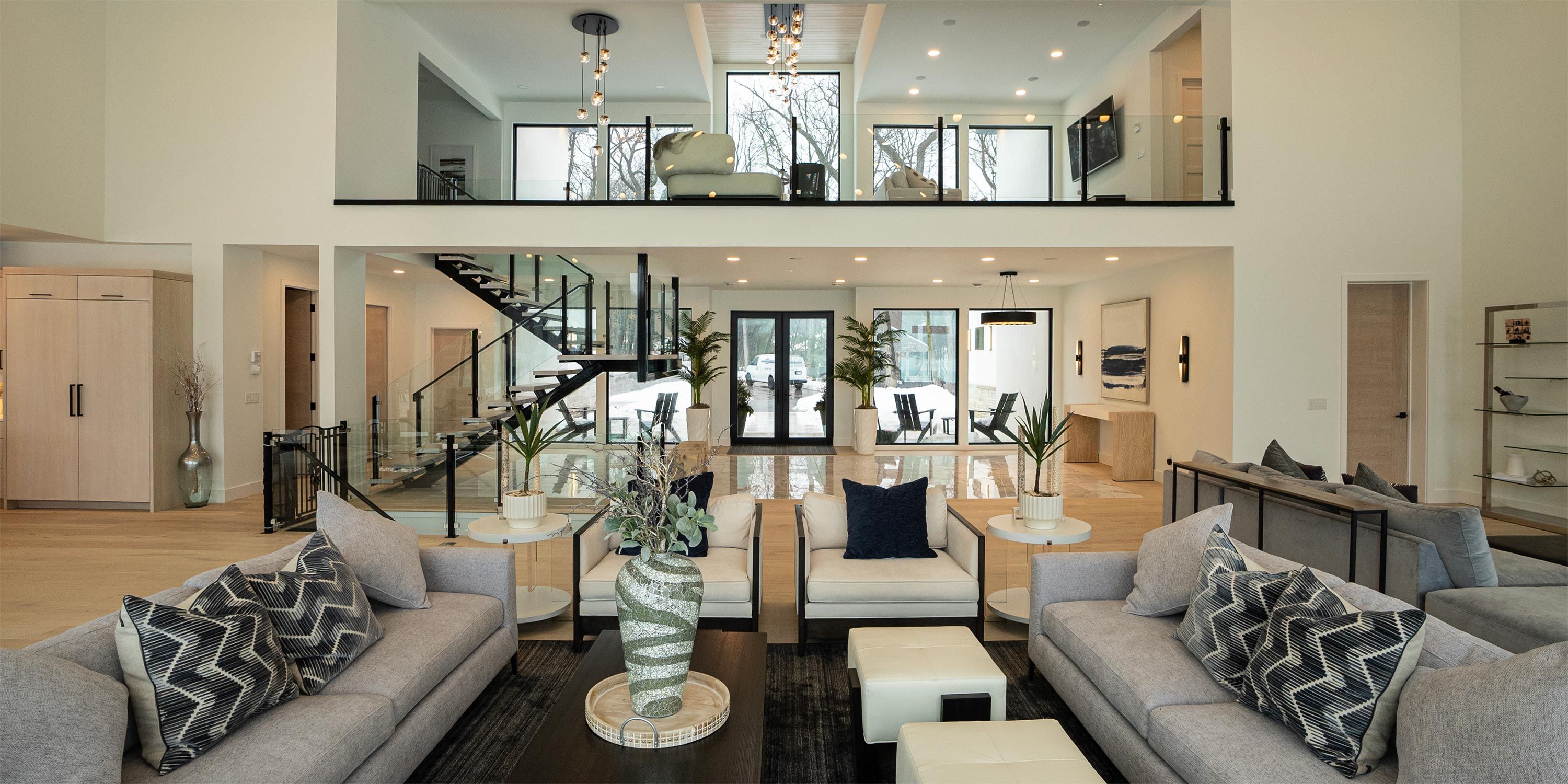 A spacious living area with a mezzanine, large windows, contemporary furnishings, and a central staircase with glass railings.