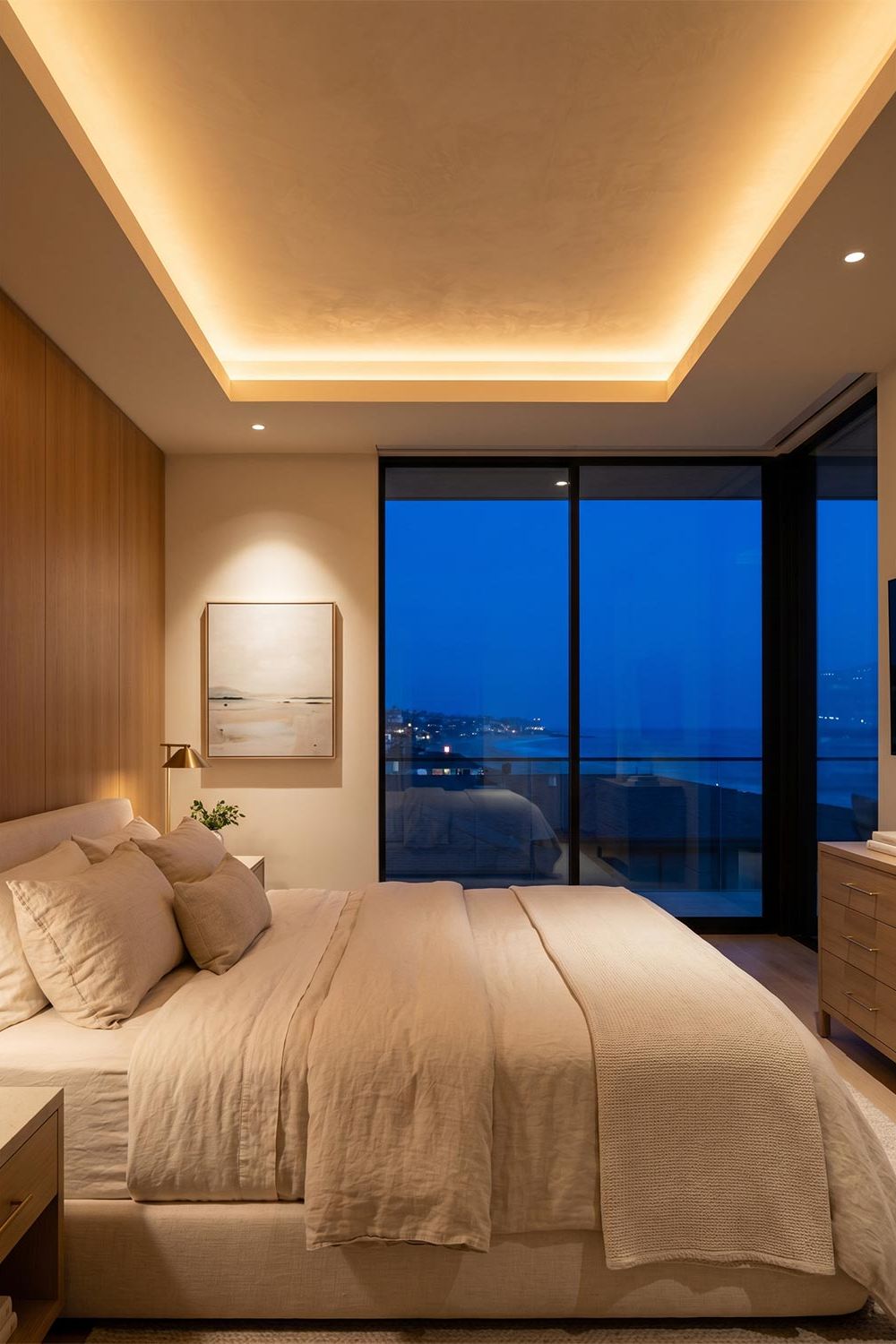 Modern bedroom with a large window overlooking the ocean at dusk and glowing ceiling light