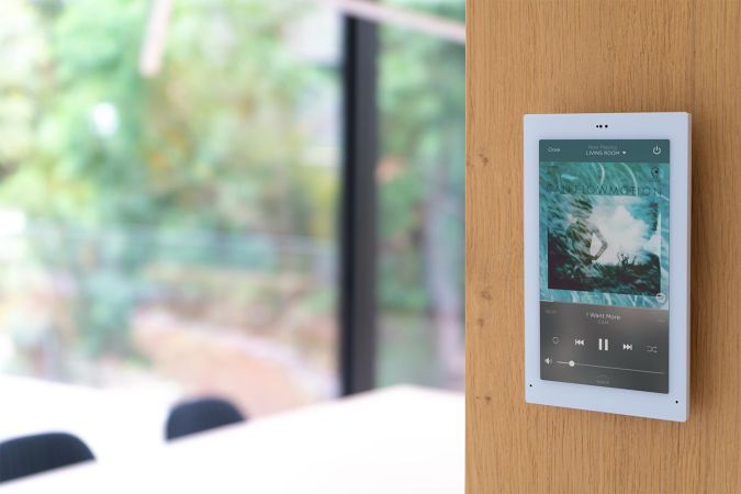 A touch screen panel mounted on a wooden wall, displaying a music playlist with a blurred view of the outdoor scenery in the background.