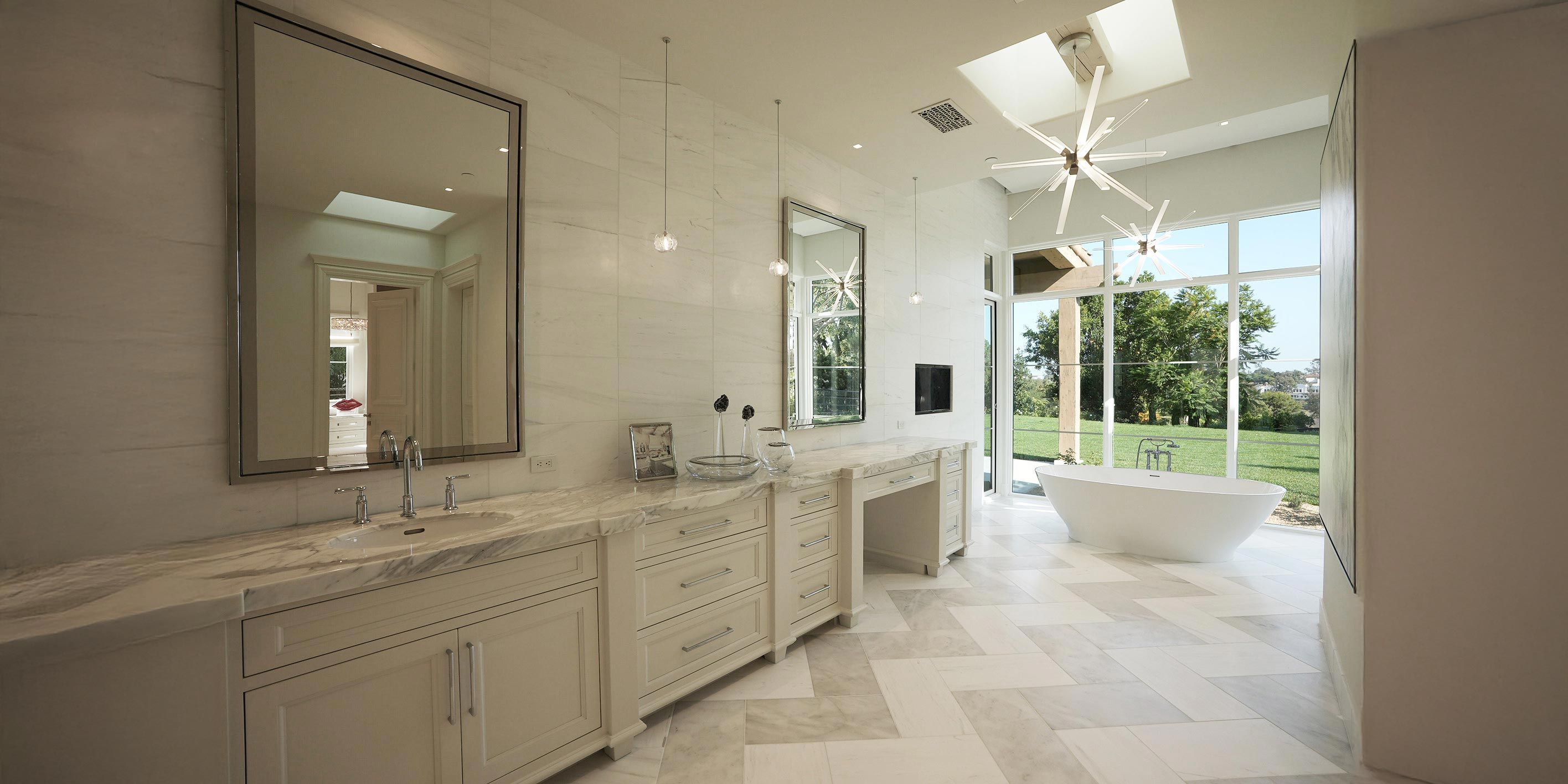 A spacious, modern bathroom featuring a long marble vanity with dual sinks, large framed mirrors, pendant lighting, a freestanding tub, and floor-to-ceiling windows overlooking a lush garden.