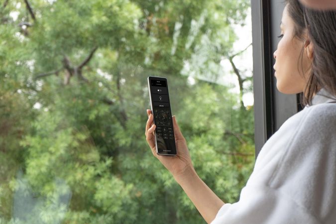 A woman holding a sleek remote control device in front of a window overlooking a lush green landscape.