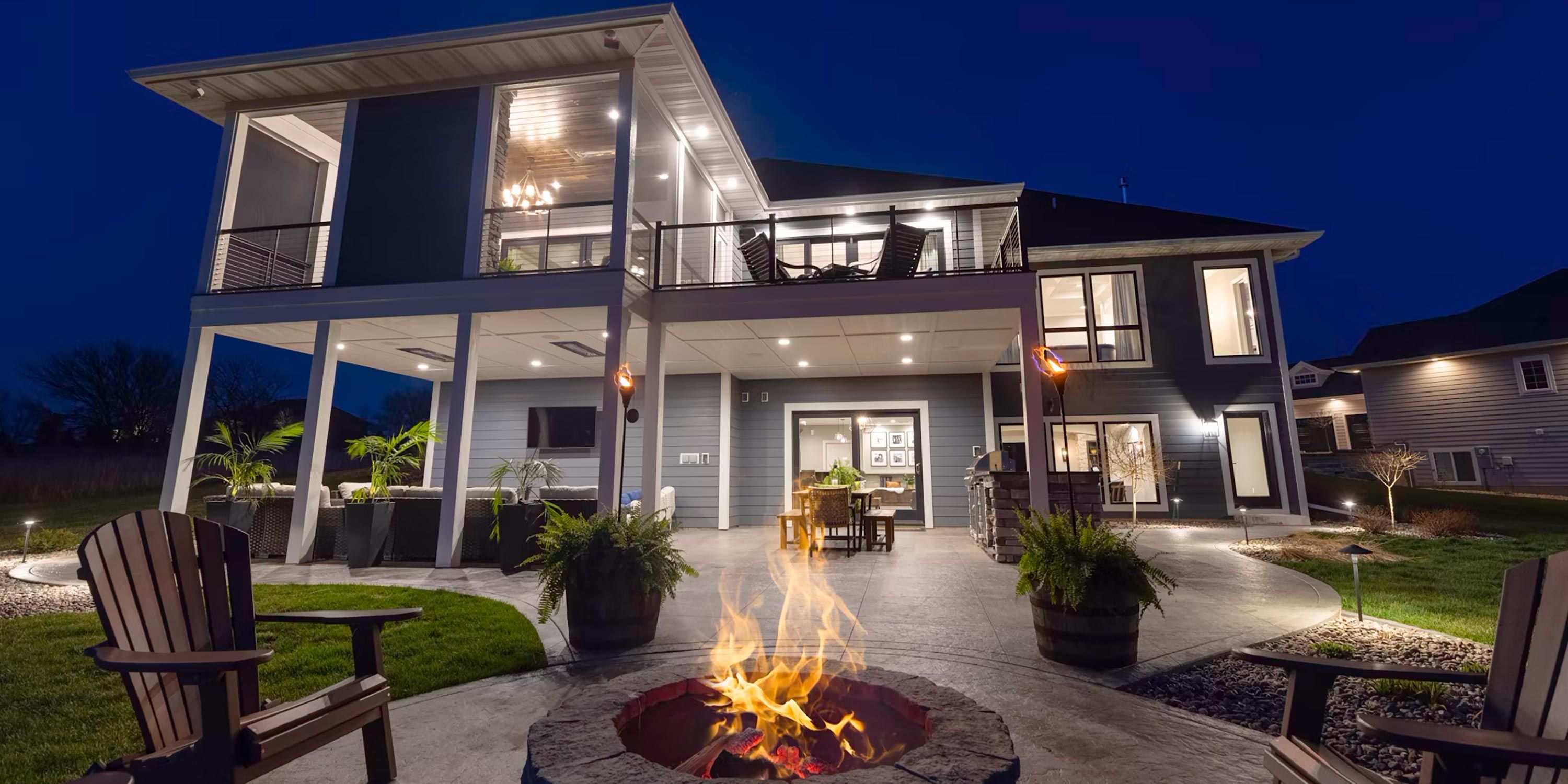 Spacious outdoor kitchen setup with a fire pit, seating, and lush landscaping under a clear night sky.